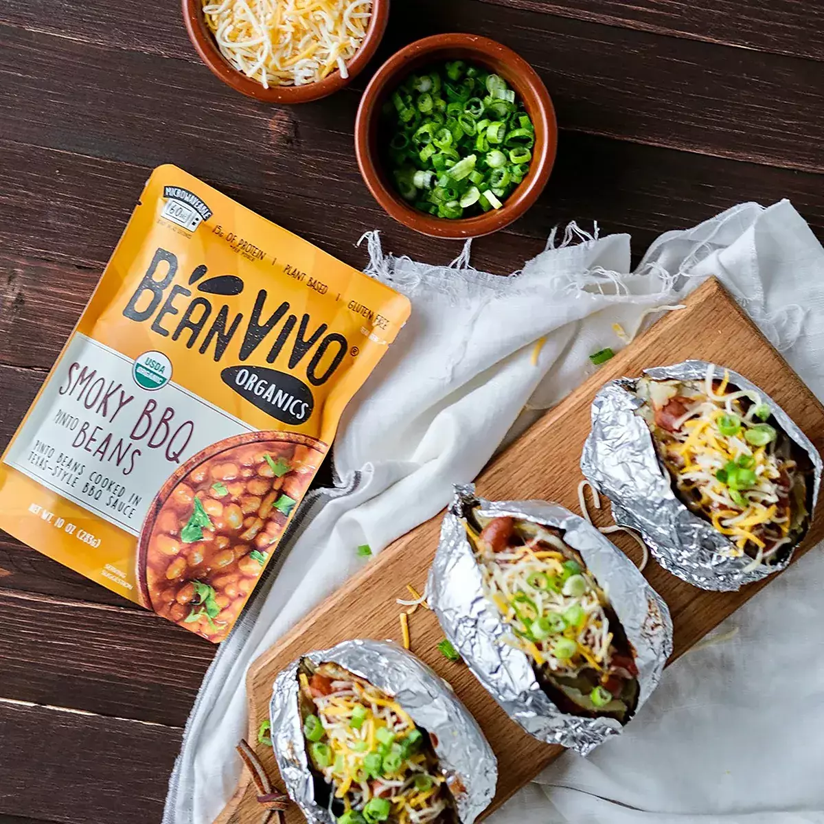 木質桌面俯視圖,右下角BeanVIVO有機煙燻燒烤斑豆包裝,上方蔥花碗乳酪絲碗,中右下方白布木托盤三份錫箔包食物含小香腸蔥花煙燻燒烤斑豆乳酪絲即食料理組合