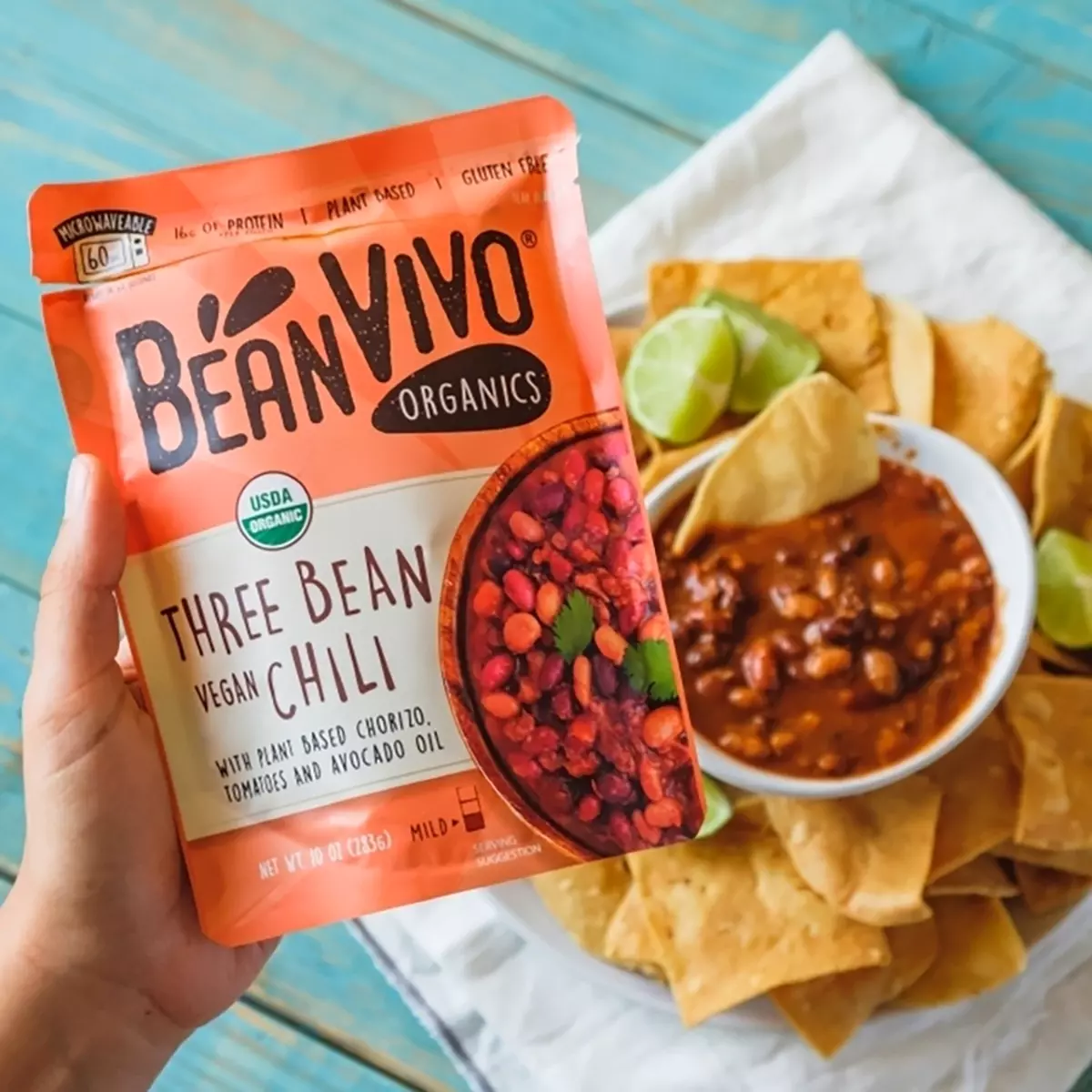 木質桌面俯視圖,左下角手拿BeanVIVO有機三豆茄汁風味包裝,中右方白布上有裝著滿滿薯片圓餐盤與一碗三豆茄汁搭配的即食料理組合