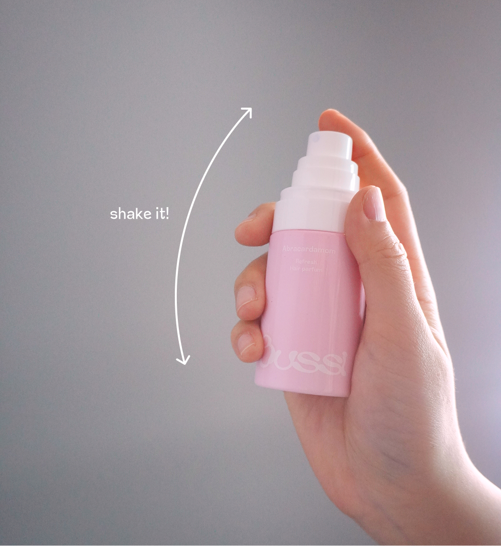 BUSSI Abracadabrum 髮香噴霧使用教學。單手持粉色瓶身噴霧，圖片附有箭頭與文字標註「shake it!」，提醒使用者在使用前先均勻搖晃產品，以獲得最佳噴灑效果