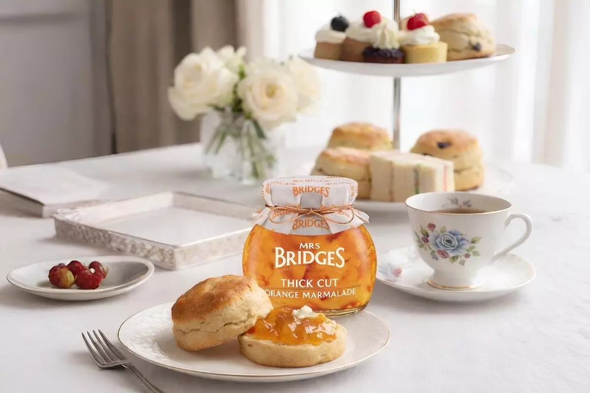 英式下午茶場景：前景盤中切開司康塗抹MRS. BRIDGES英橋夫人厚切柑橘果醬與奶油；後方果醬產品瓶，右側骨瓷紅茶杯，右後點心塔堆疊小蛋糕司康，左後玻璃瓶插白玫瑰，展現經典英倫風華。
