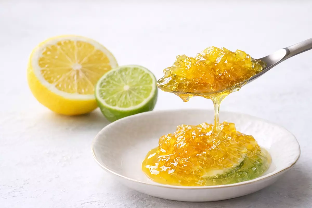 檸檬萊姆果醬微距攝影，展現柑橘果肉與細緻果皮。一匙半透明果醬懸浮滴落，突顯濃稠質感與新鮮酸度。左側配半顆多汁檸檬與翠綠萊姆，漸層金黃綠色澤自然。