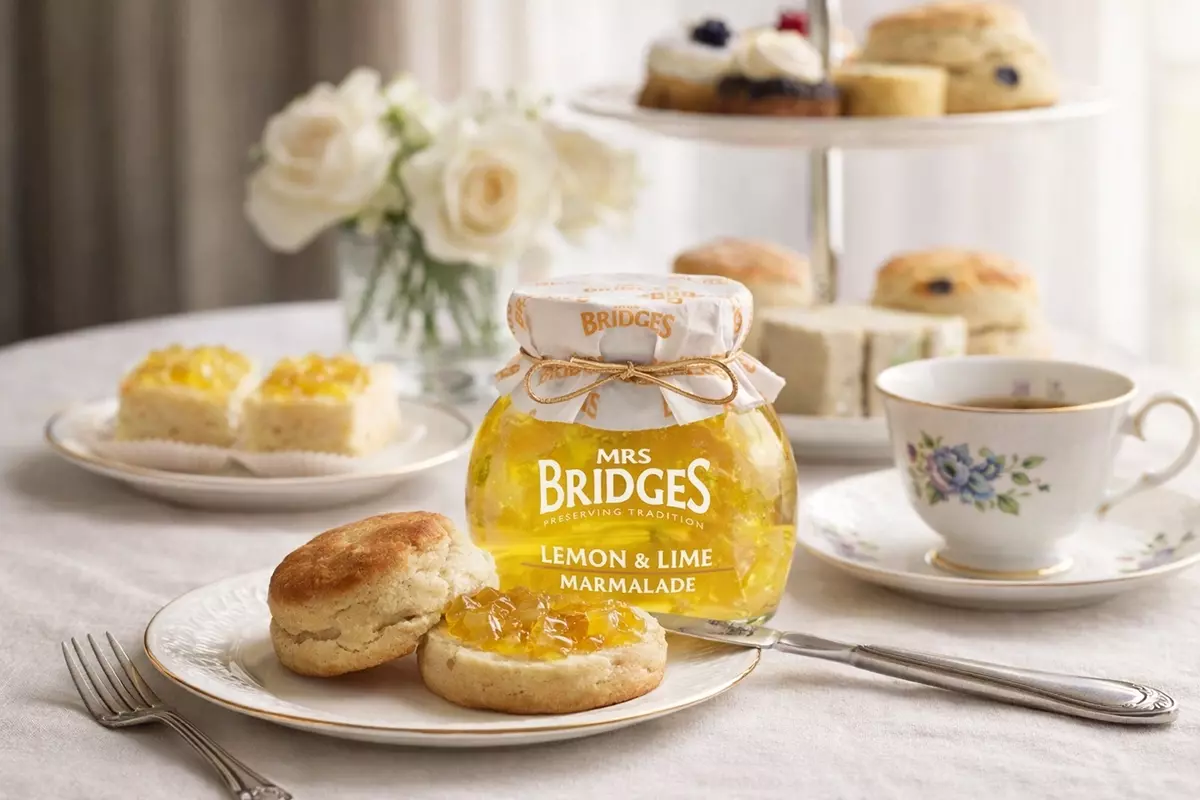 英式下午茶場景：前景盤中切開司康塗抹MRS. BRIDGES英橋夫人檸檬萊姆果醬；後方果醬產品瓶，右側骨瓷紅茶杯，右後點心塔堆疊小蛋糕司康，左後玻璃瓶插白玫瑰與塗抹檸檬萊姆果醬小蛋糕。