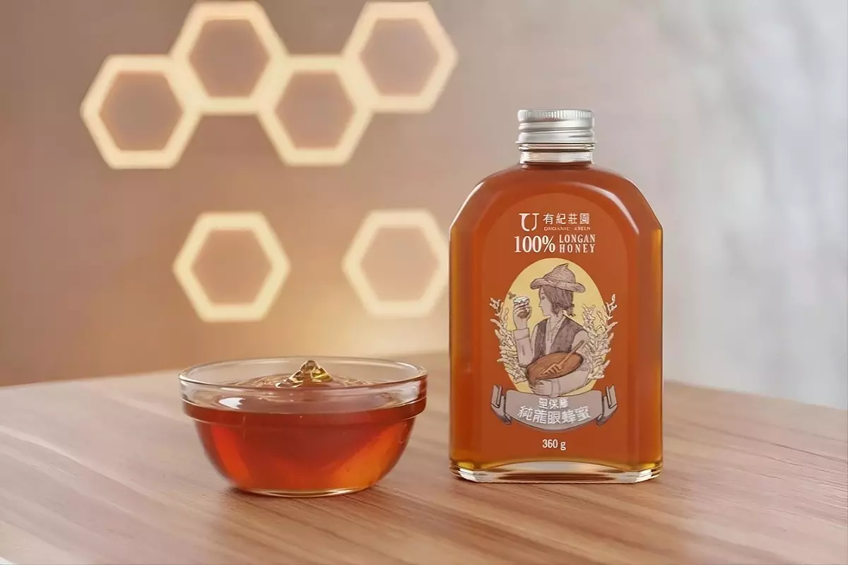 玻璃碗中的濃琥珀色蜂蜜放置在溫暖木質桌面上，蜂蜜厚實光亮、微微流動，玻璃折射自然光澤。旁邊擺放聖保羅 100% 純龍眼蜂蜜瓶，背景為柔焦蜂巢景觀，營造溫馨、高質感、誘人的氛圍。