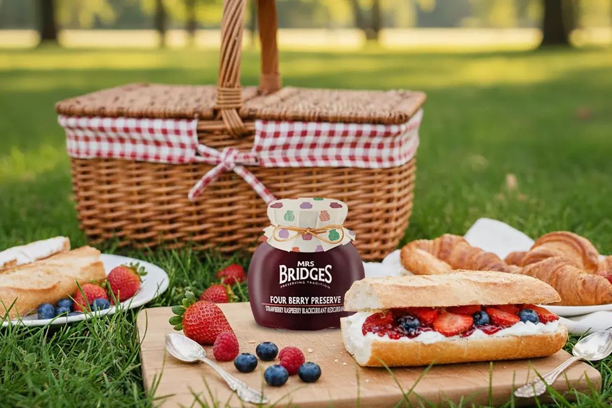 優雅野餐場景置於綠草地上，MRS. BRIDGES 英橋夫人莓果四重奏綜合果醬罐，內含閃耀莓果果醬，旁邊法式麵包三明治展現果塊，質樸籃子相伴，柔和自然陽光。