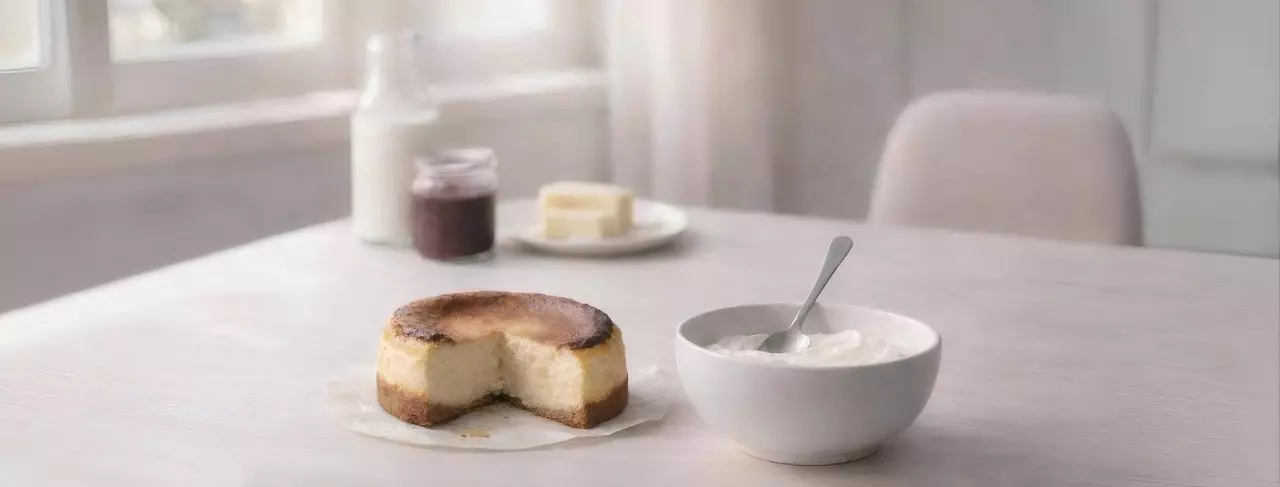 明亮木質餐桌上的溫馨早餐，切半巴斯克蛋糕、挖過優格、牛奶起司果醬瓶，自然柔和光線