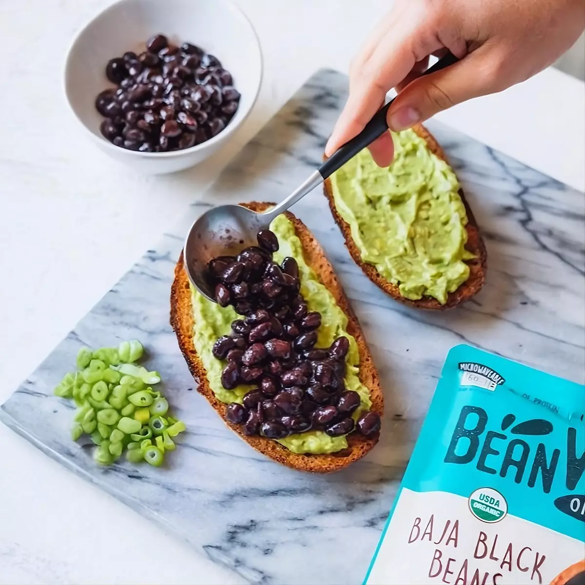 大理石桌面俯視圖，右下角BeanVIVO有機巴哈黑豆包裝，中右方手拿勺子舀有機黑豆鋪在抹有醬料的麵包上即食料理組合