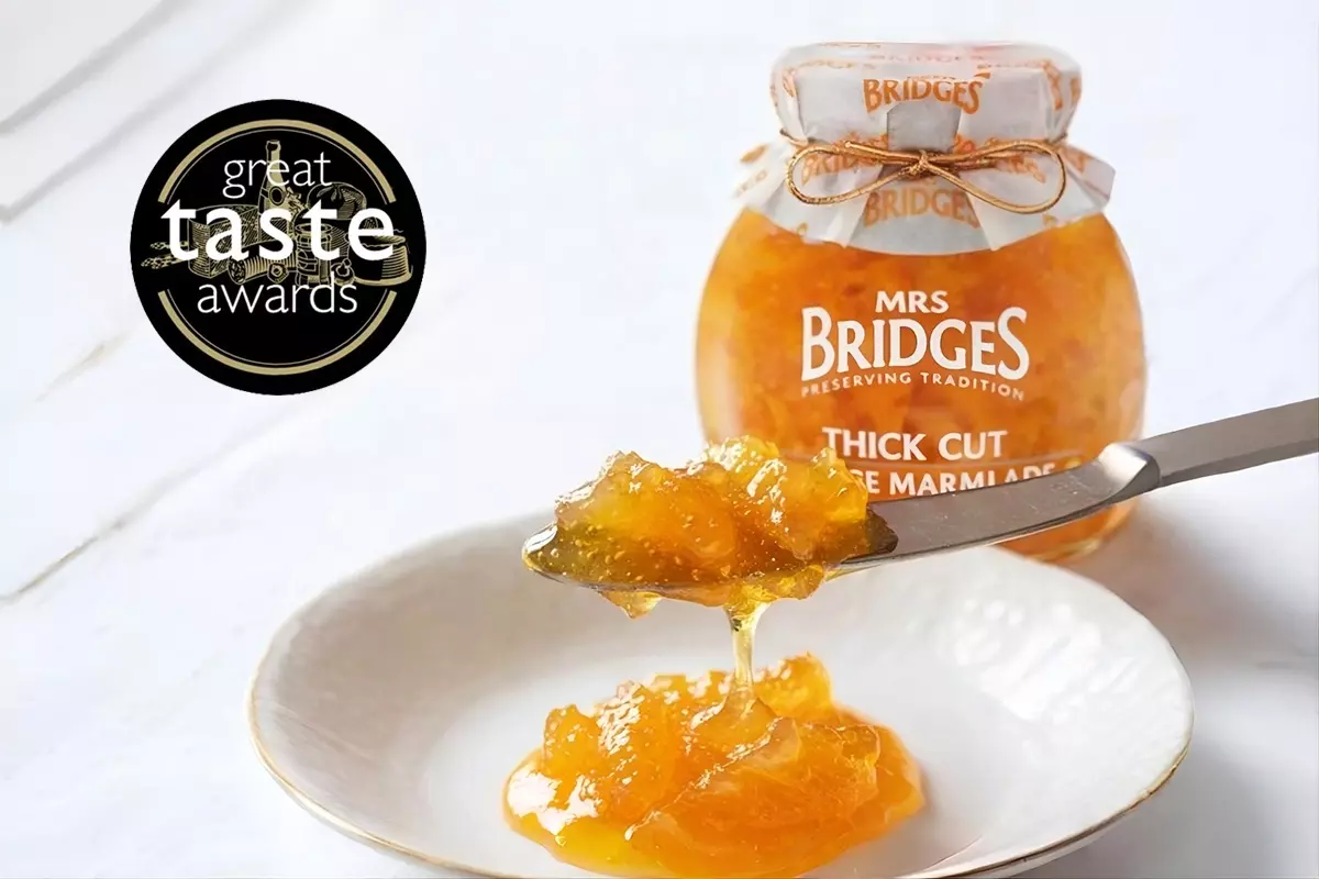 白色木質桌面上擺放一盤金黃柑橘果醬，果醬抹刀正舀起一小匙香濃果醬；盤子右後方陳列MRS. BRIDGES英橋夫人厚切柑橘果醬產品瓶，左上方飾以Great Taste Awards獲獎徽章，展現英式經典風味。