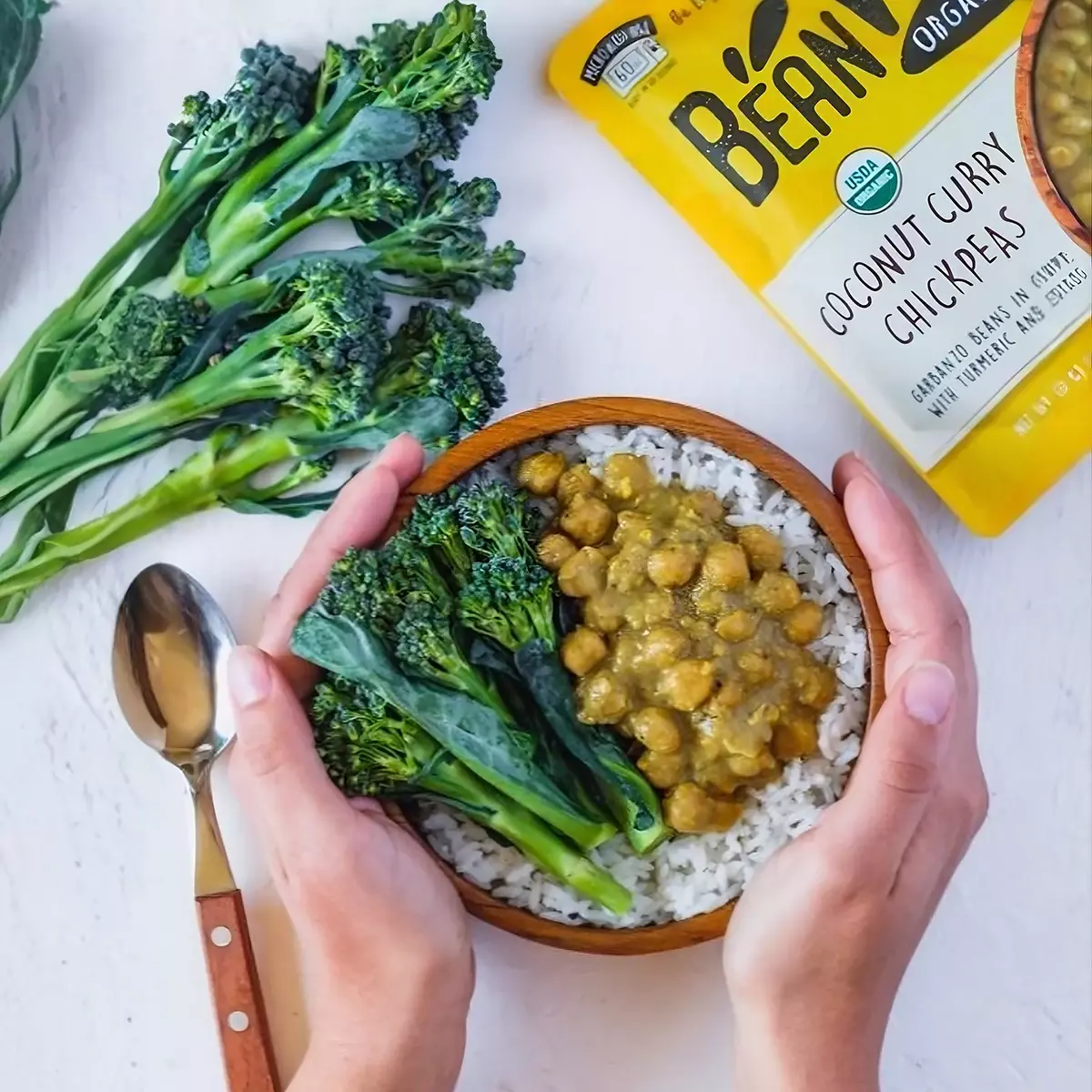 灰白色木質桌面俯視圖，左上角BeanVIVO有機椰奶咖哩鷹嘴豆包裝，中間手捧一碗米飯淋上有機椰奶咖哩鷹嘴豆，搭配花椰菜即食料理組合
